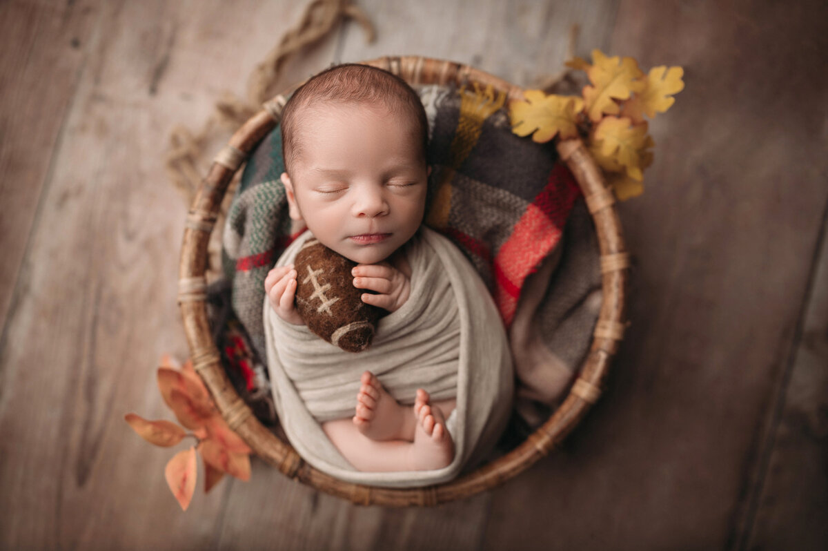 two week old newborn sleeping baby boy  wrapped in a cream stretch wrap and holding a tiny felted football posed in round wooden bowl over a plaid blanket surrounded by yellow and orange fall leaves on top of wooden floor