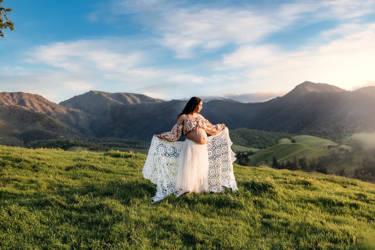 pregnant woman in a white 2 piece maternity gown. she holds a knit blanket and mt diablo is behind her
