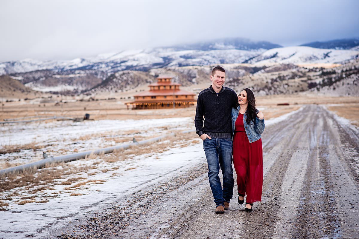 Girl wearing full-length red dress and jean jacket walking with her fiance down a gravel road at Headwaters Ranch with a circular barn behind them and snow-capped mountains