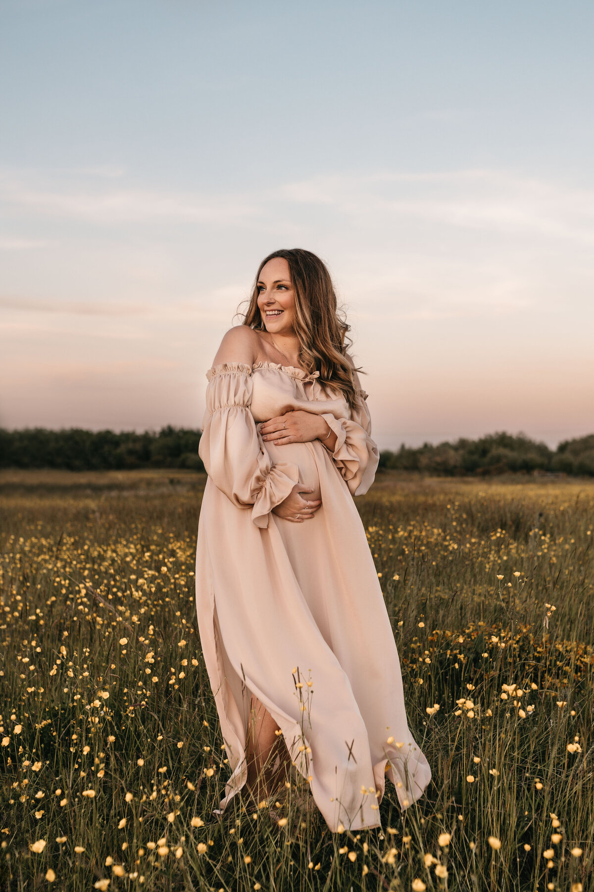 Photo of a pregnant woman holding her bump and standing in a field of flowers at sunsetAC