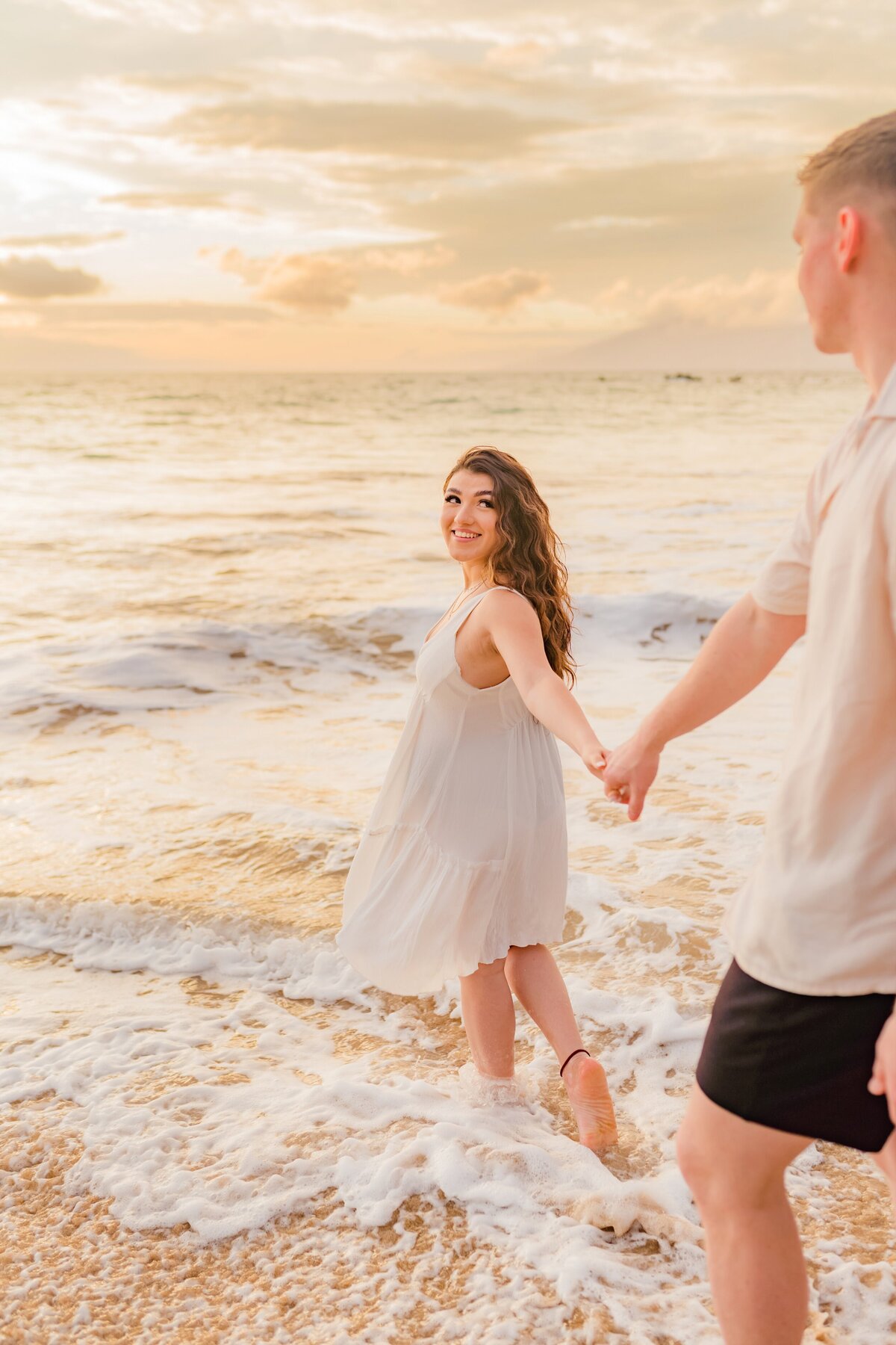 Brunette woman wearing a white dress leads her boyfriend into the water on the beach in Hawaii