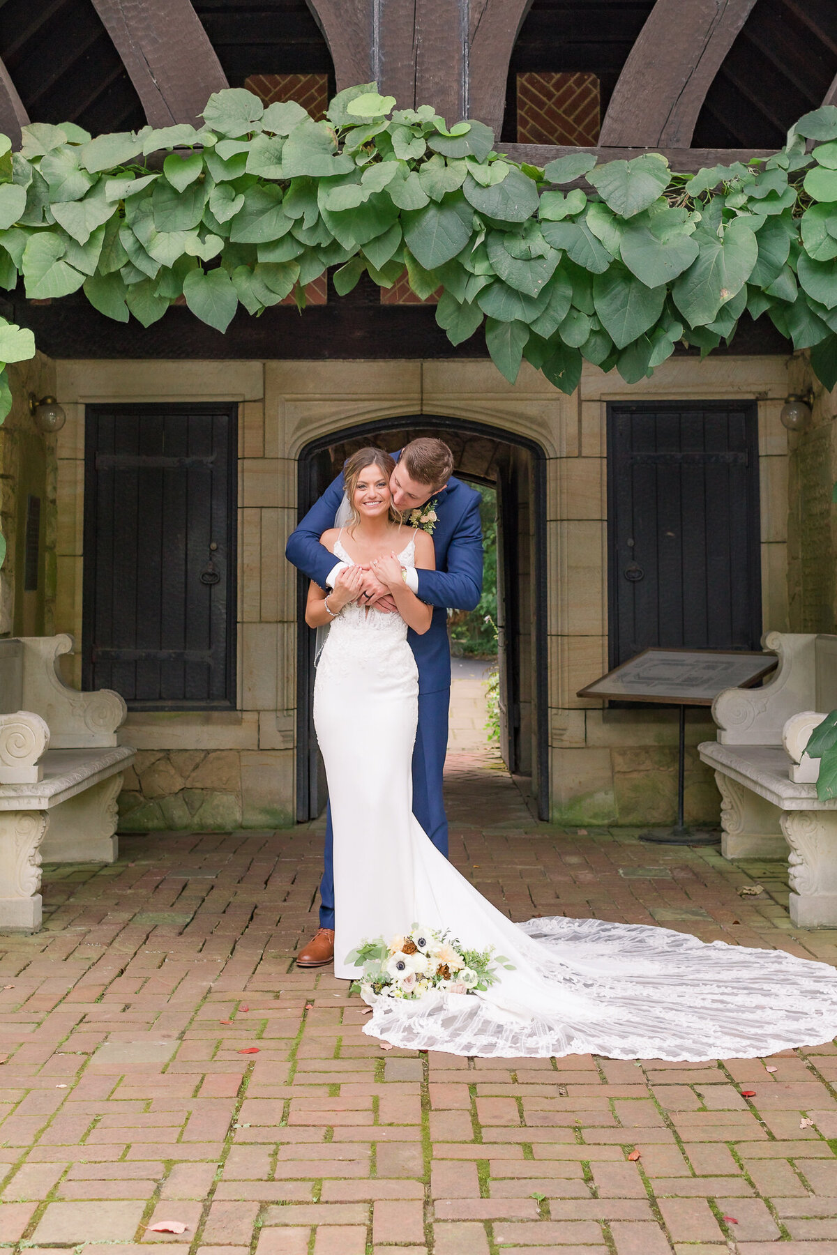GROOM IN NAVY SUIT SOFTLY EMBRACES HIS NEW BRIDE UNDER AN  ARBOR OF DUTCHMAN'S PIPE VINES