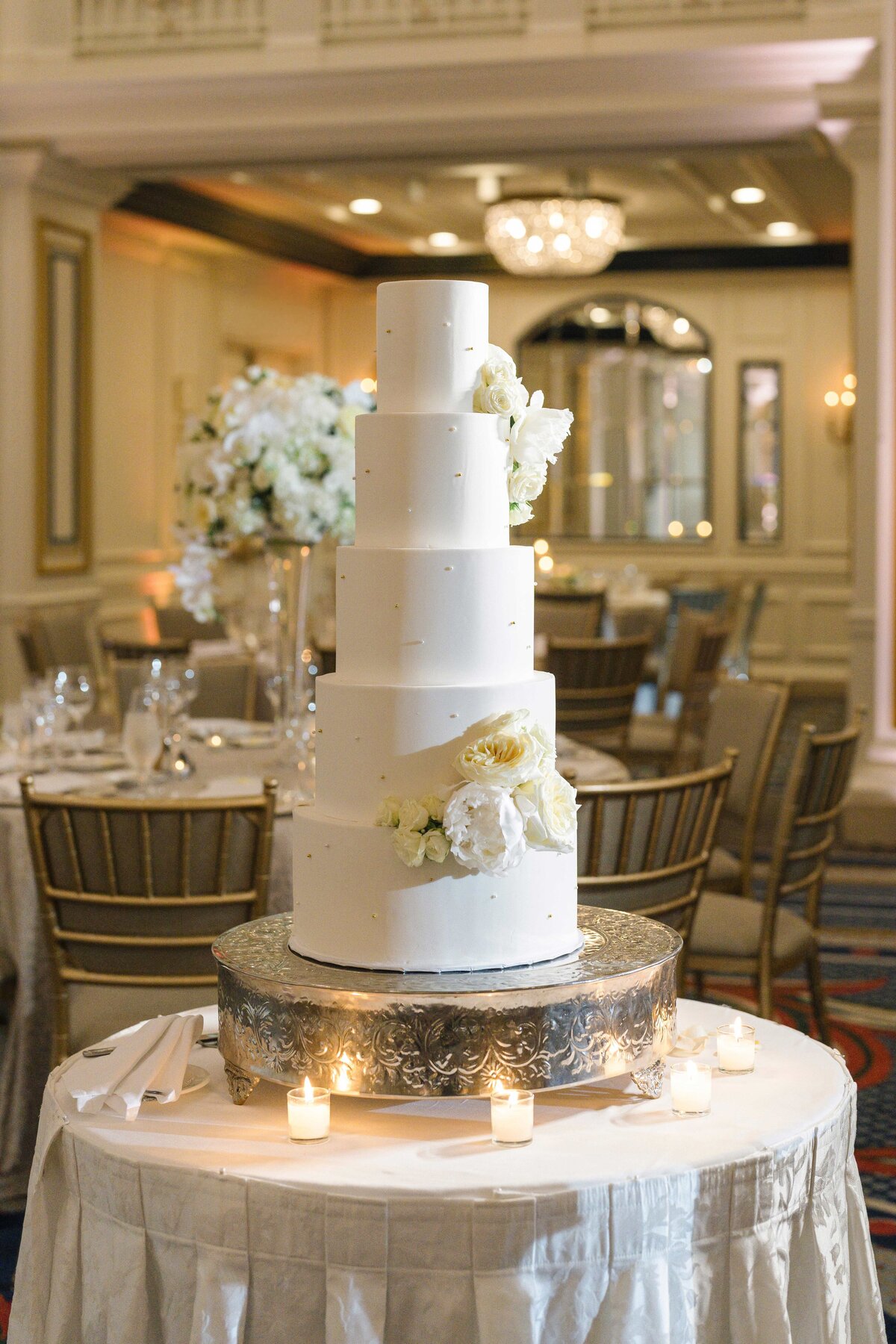 A four-tier white wedding cake adorned with white flowers is on an ornate silver stand. The table is surrounded by small candles. The background features an elegantly decorated event space with tables and chairs.
