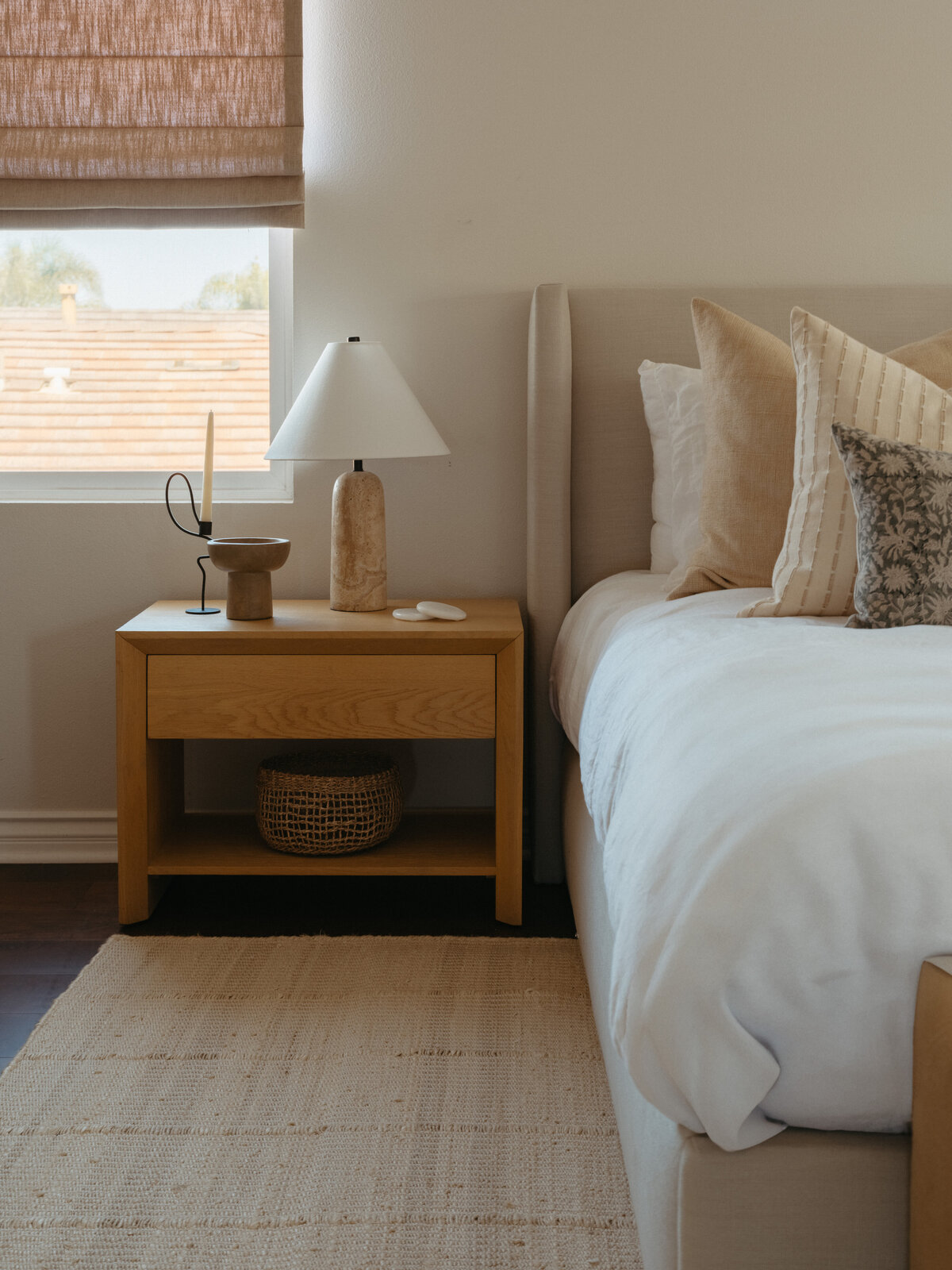 Organic modern bedroom design by Natalie Barnas Interiors, a boutique interior design studio serving Carlsbad, Encinitas, Del Mar, La Jolla, Solana Beach, and Coronado, California. This serene corner vignette features a custom upholstered platform bed layered with neutral linen and textured accent pillows, a natural oak nightstand styled with a travertine lamp, minimalist candleholder, and ceramic decor, all grounded by a handwoven jute rug and soft Roman shades. A refined example of California coastal interior design blending warmth, texture, and minimalist luxury.