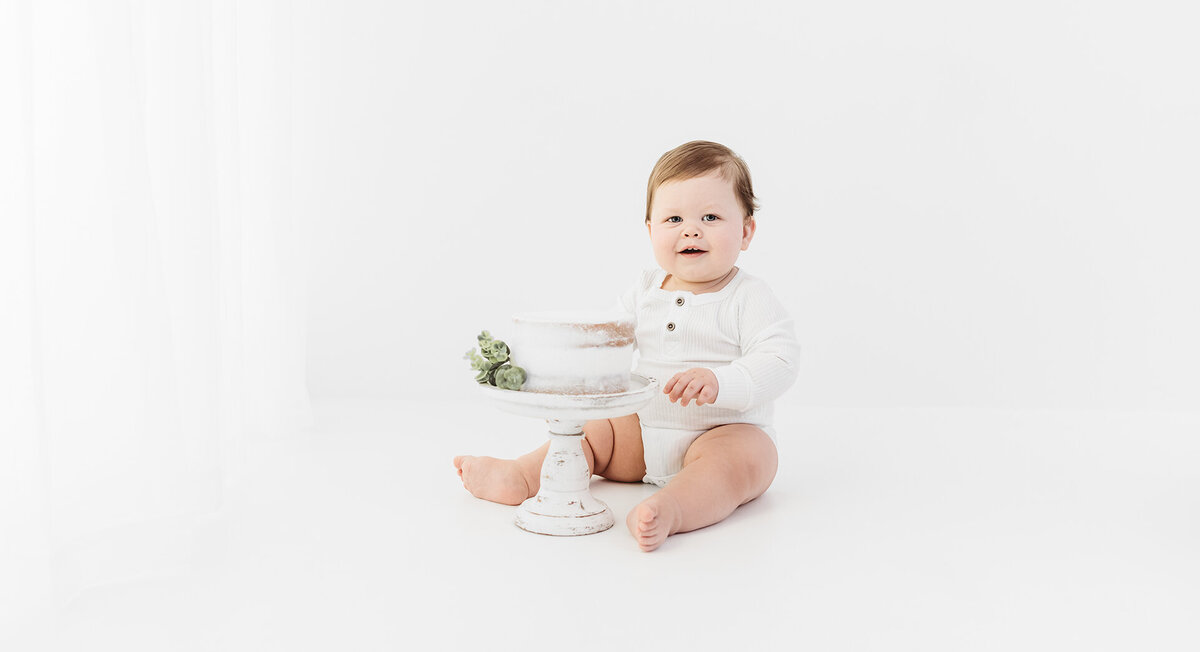 The photo shows an adorable one-year-old baby boy celebrating his first birthday with a cake smash photo session. He is dressed in a white onesie, matching white backdrop, and a white cake. The baby looks overjoyed as he happily enjoys his birthday cake, creating a memorable moment captured by photographer Bri Sullivan in a clean, pure, and classic style.