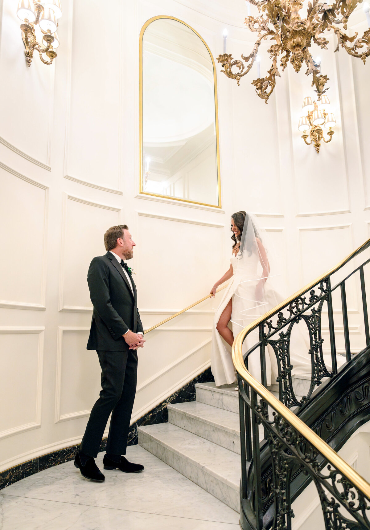 A bride in a white dress and veil descends a grand staircase, smiling at a man in a suit waiting at the bottom. The elegant setting features ornate lighting and a large mirror.