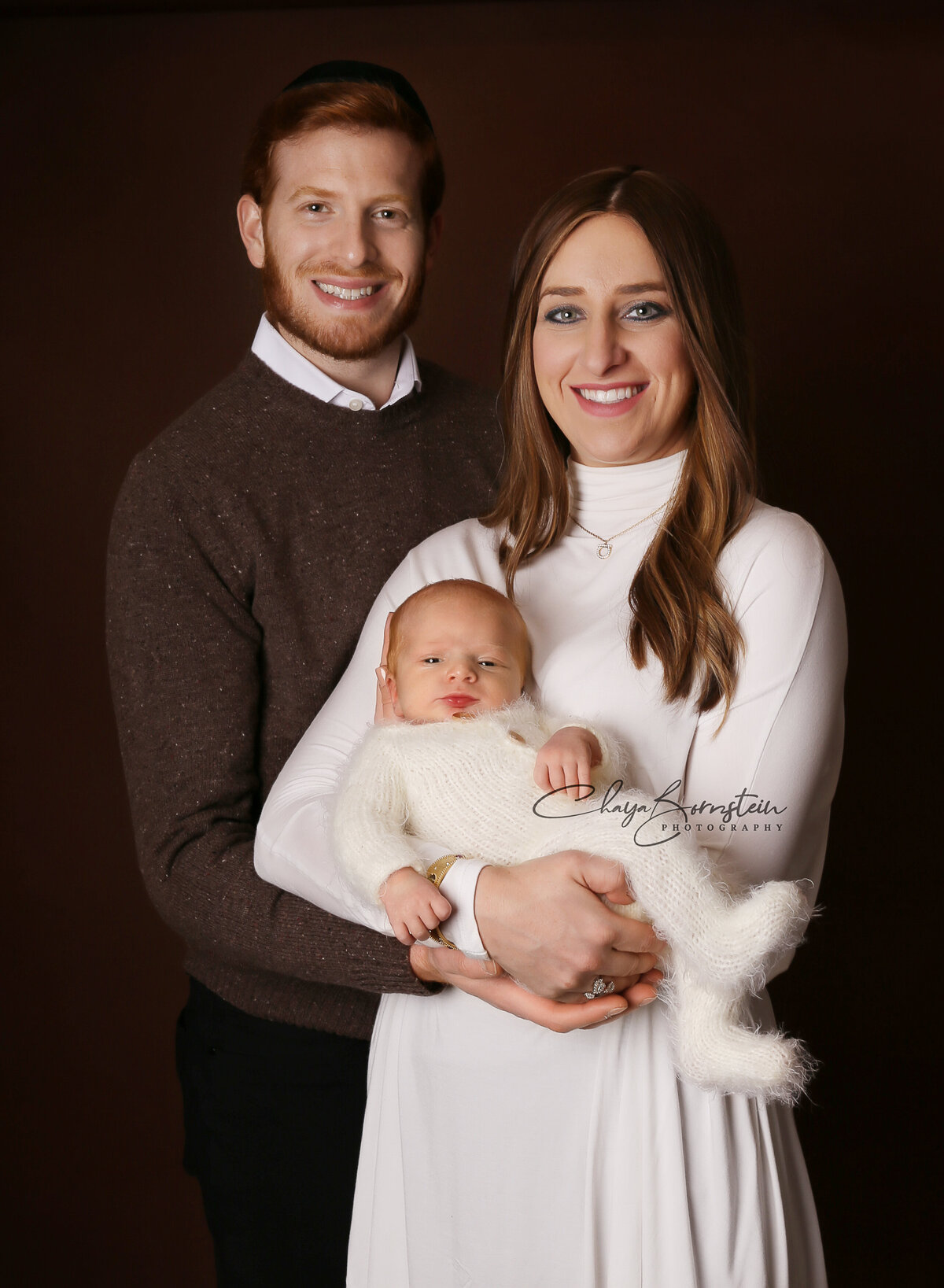 Mom and dad pose for a newborn photoshoot with their newborn baby. Mom and dad are smiling at the camera and baby is awake. Captured by top Brooklyn newborn photographer Chaya Bornstein