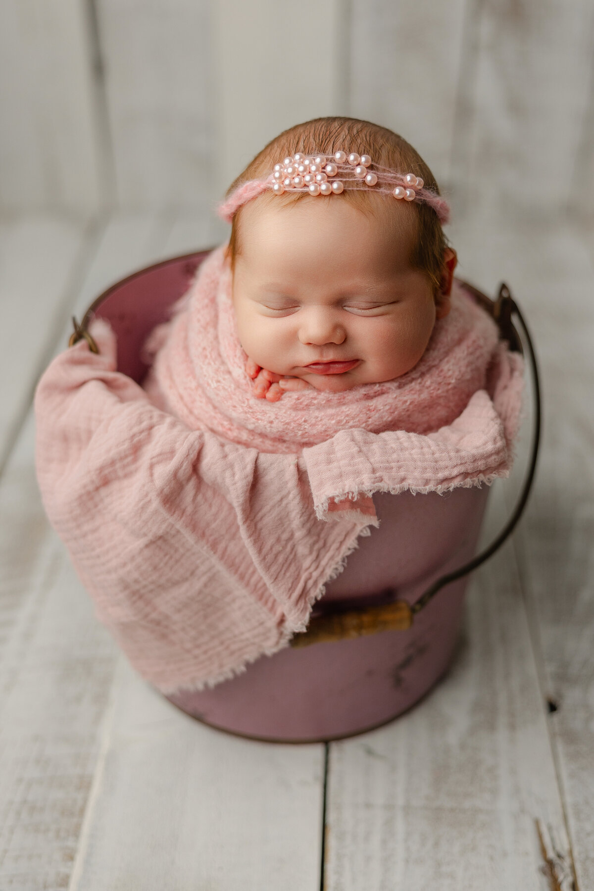 Smiling newborn baby girl swaddled in pink with a pearl headband, posed in a rose-colored bucket during her studio session. Captured by Inland Empire newborn photographer Kaitlyn Dawn Photography.
