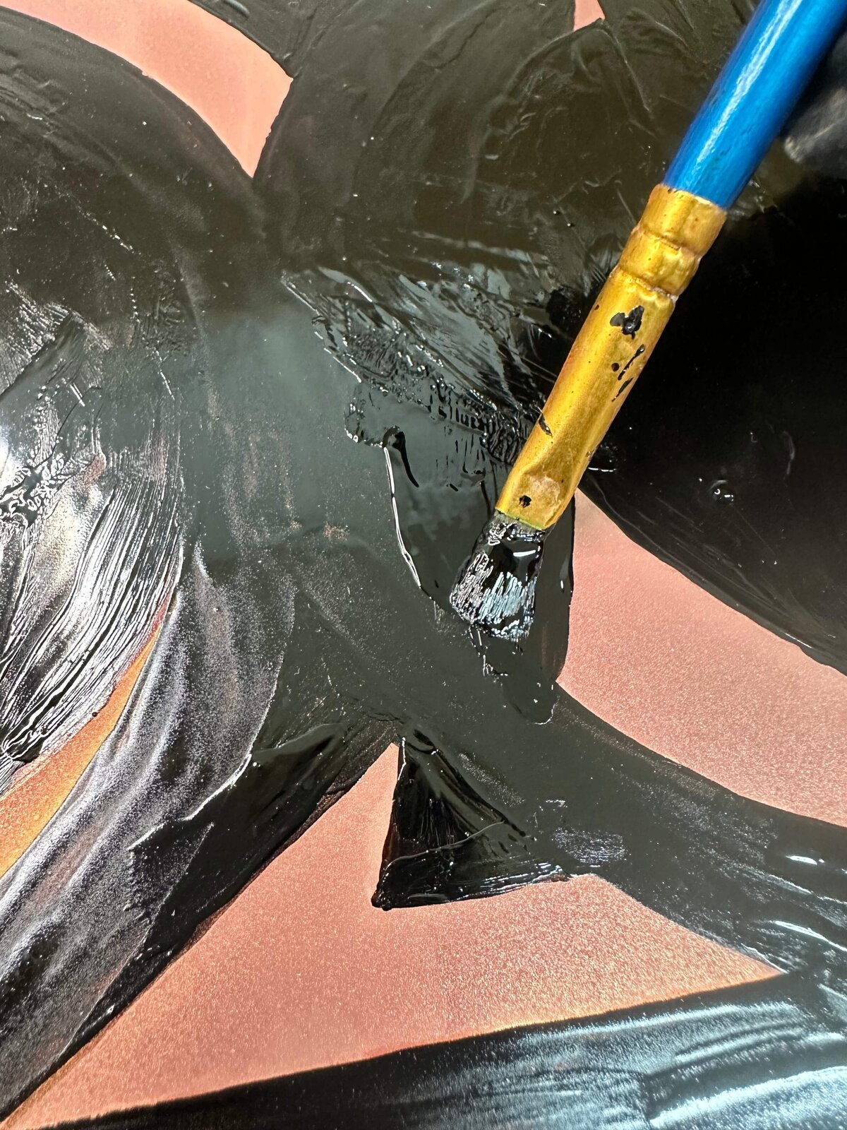 A close-up of a brush applying black asphaltum to a copper plate, demonstrating fine control in blocking out areas for step etching.