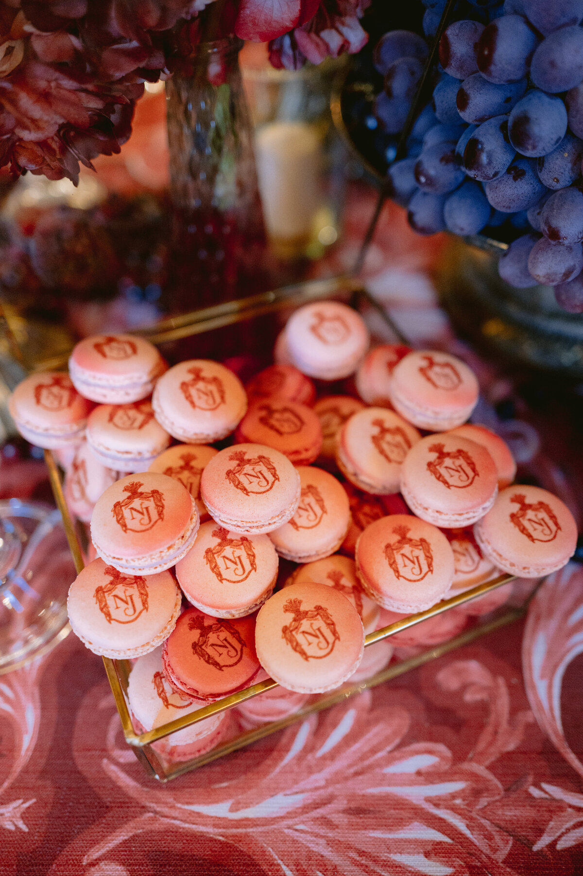 The photo was taken at the breathtaking Shangri La Hotel and Ritz Paris Hotel in Paris, France, capturing a stunning display of a glass tray filled with pink and peach-colored macarons. These delicate treats were expertly decorated with an intricate crest design, exuding a touch of sophistication. The background of the image showcases a charming setting with a floral tablecloth, clusters of purple grapes, and blurred flowers, creating a vibrant and elegant ambiance that enhances the overall aesthetic. The photo was skillfully captured by the talented photographer Matteo Coltro, with Videographers Zen Film Works also involved in documenting the scene in a captivating manner.