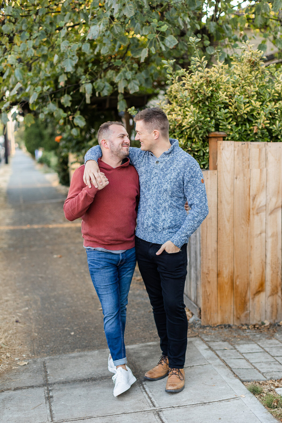 Two people walking arm in arm on a sidewalk beside a wooden fence and trees