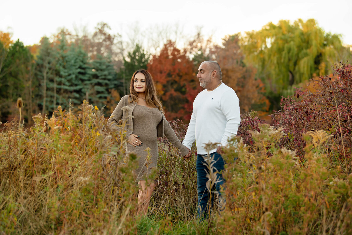 husband and wife holding hands in a field for their fall maternity photos in Burlington