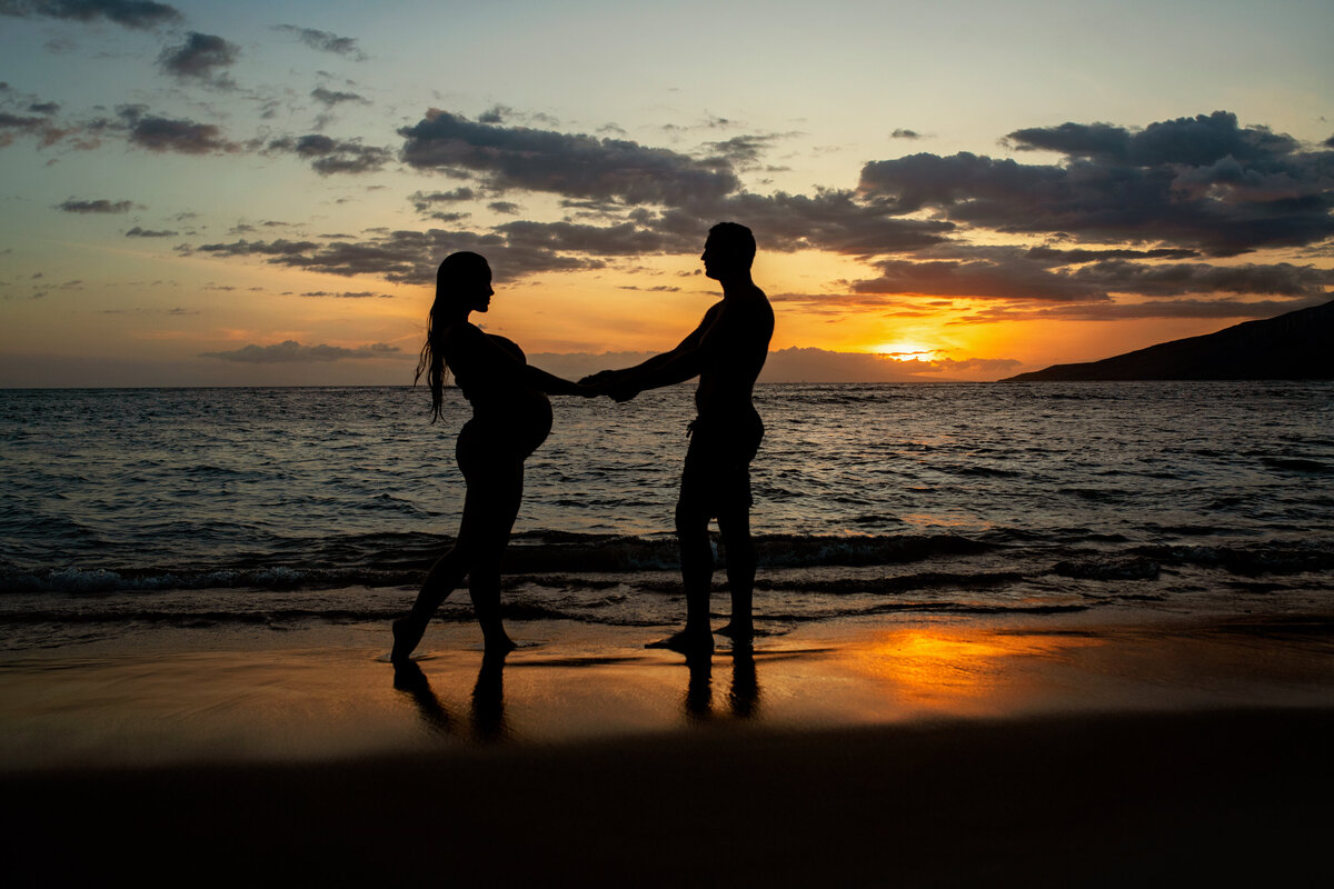 Soon to be parents holding hands with baby bump showing at the beach during sunset in Maui