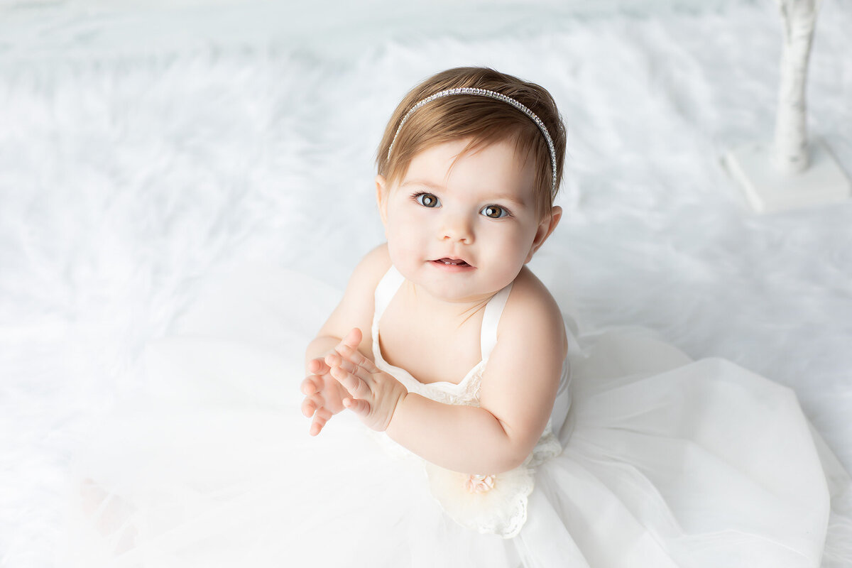 Dallas baby photographer captures a milestone portrait of a little girl in a white gown and jeweled headband, clapping softly on a fluffy snow-white backdrop.
