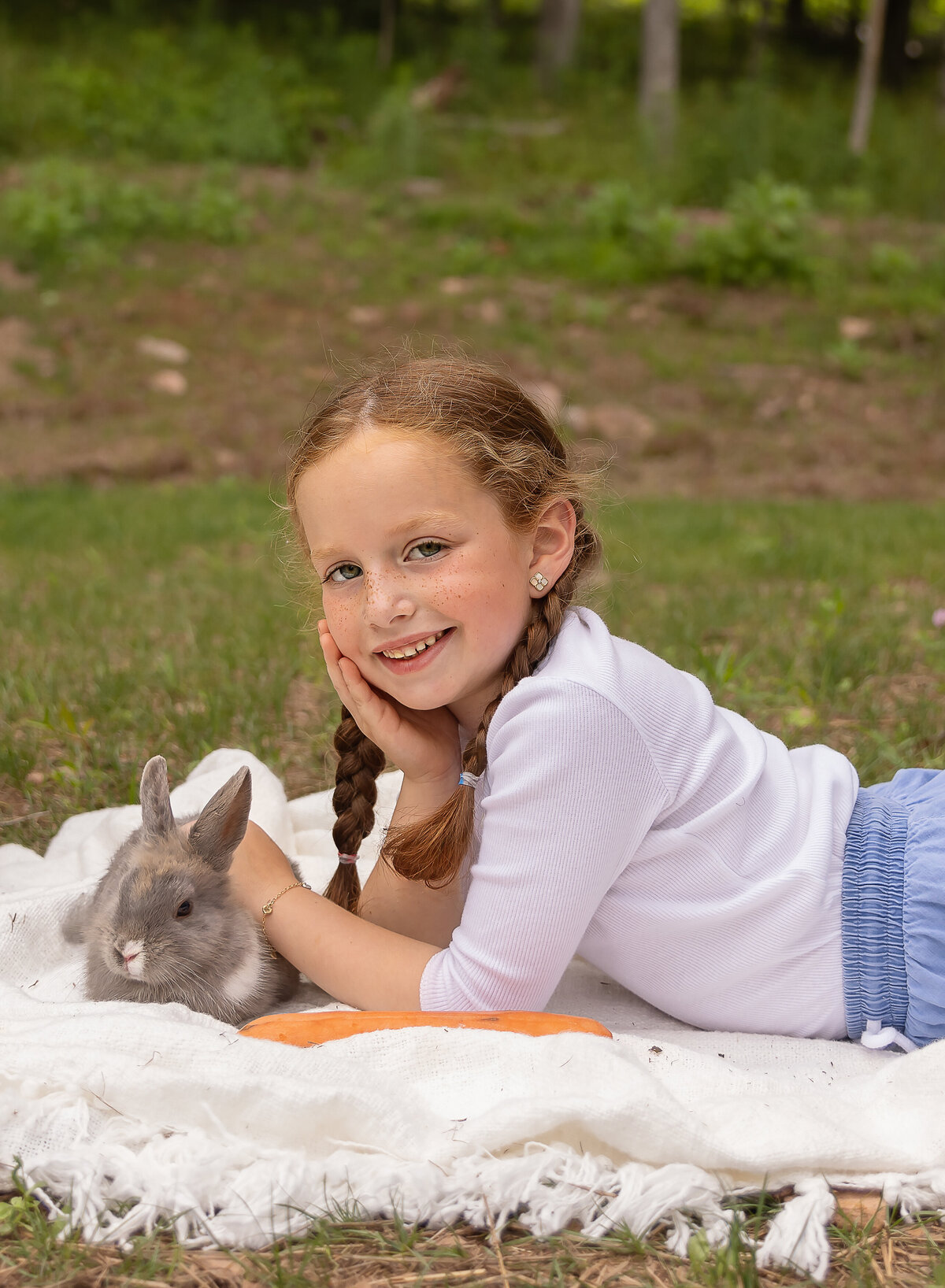Little girl is laying on a blanket with a bunny for a brooklyn, ny photoshoot. She is looking over her shoulder and smiling at the camera. Captured by brooklyn family photographer Chaya Bornstein