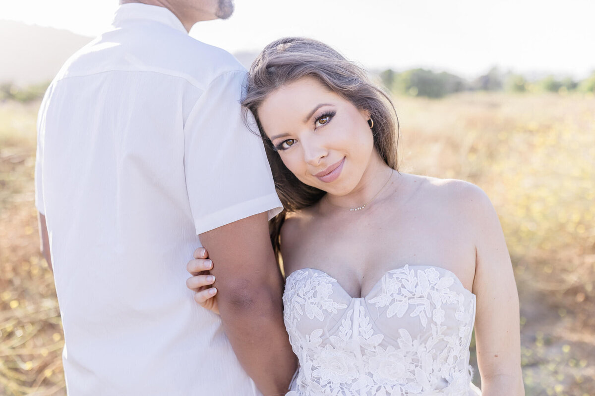 A woman in a strapless lace dress stands smiling next to a man in a white shirt in an outdoor setting with blurred trees and grass in the background. The scene is bright and sunny.