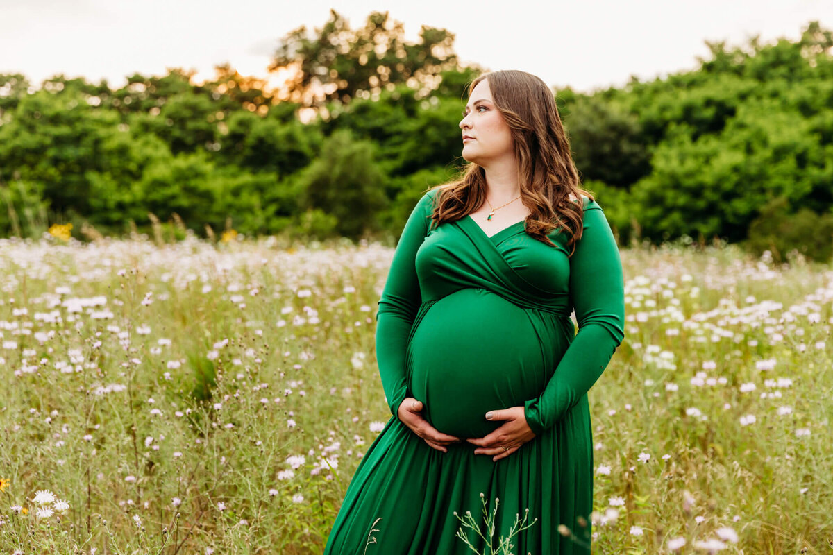 stunning pregnant mom in a flowy green dress  holding onto her baby bump as she looks out into the wildflowers captured by Ashley Kalbus Photography