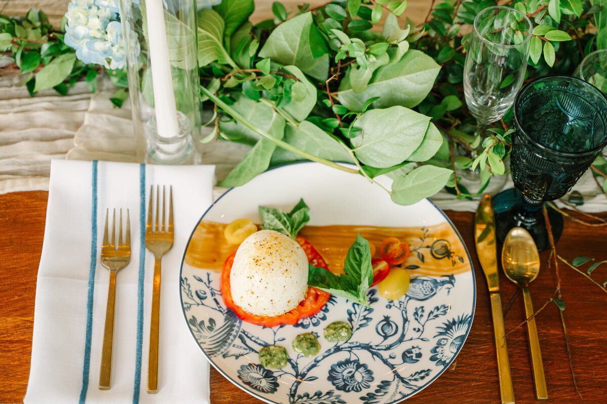 a wooden table set with gold flatware blue stemware and a floral blue and white china plate with a tomato caprese salad with torn basil and painted balsamic reduction at Willowbrook wedding venue