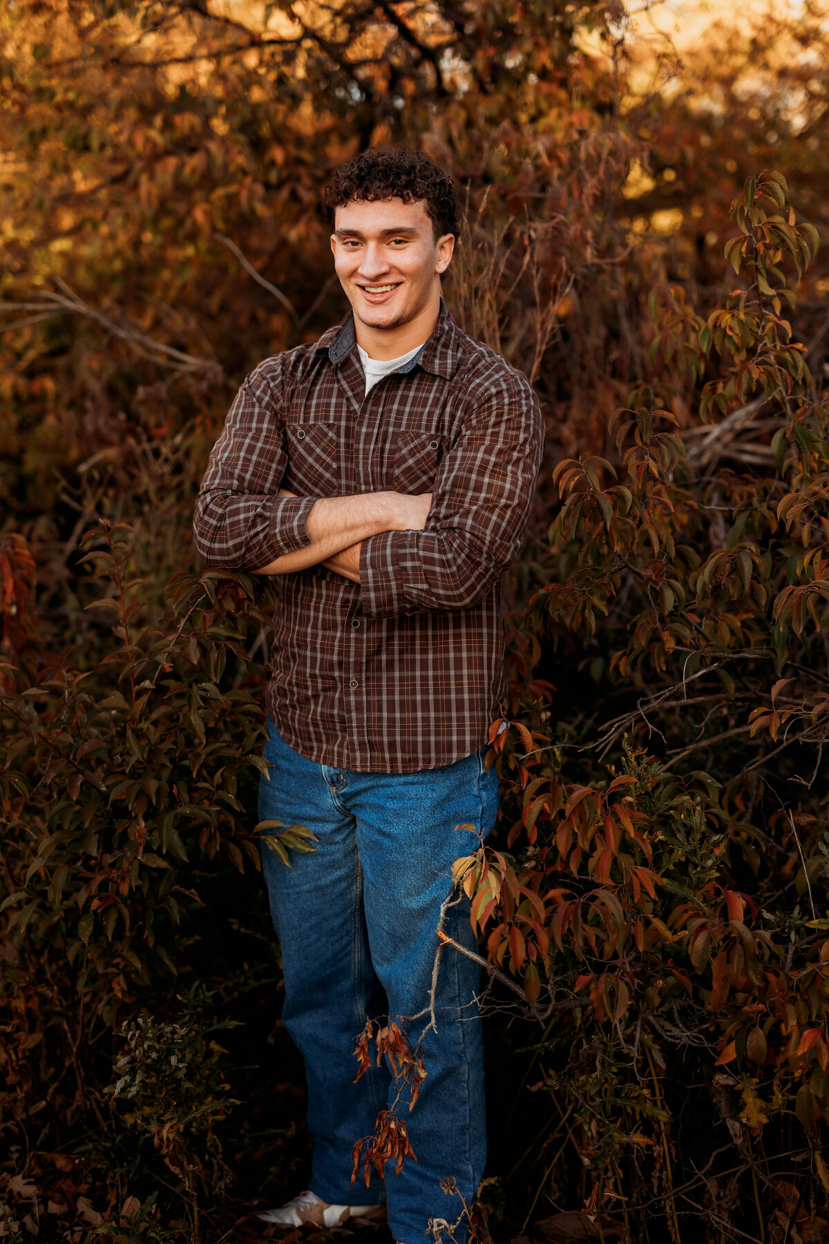 Full body image of a high school senior boy in jeans and a plaid button up standing in front of fall colored bush