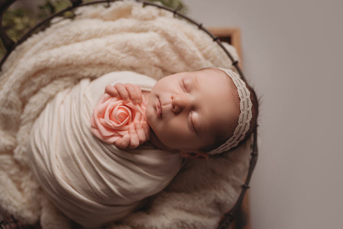 Sleeping newborn girl dressed in white swaddle and lace headband holding a rose in her hands and laying on faux floor