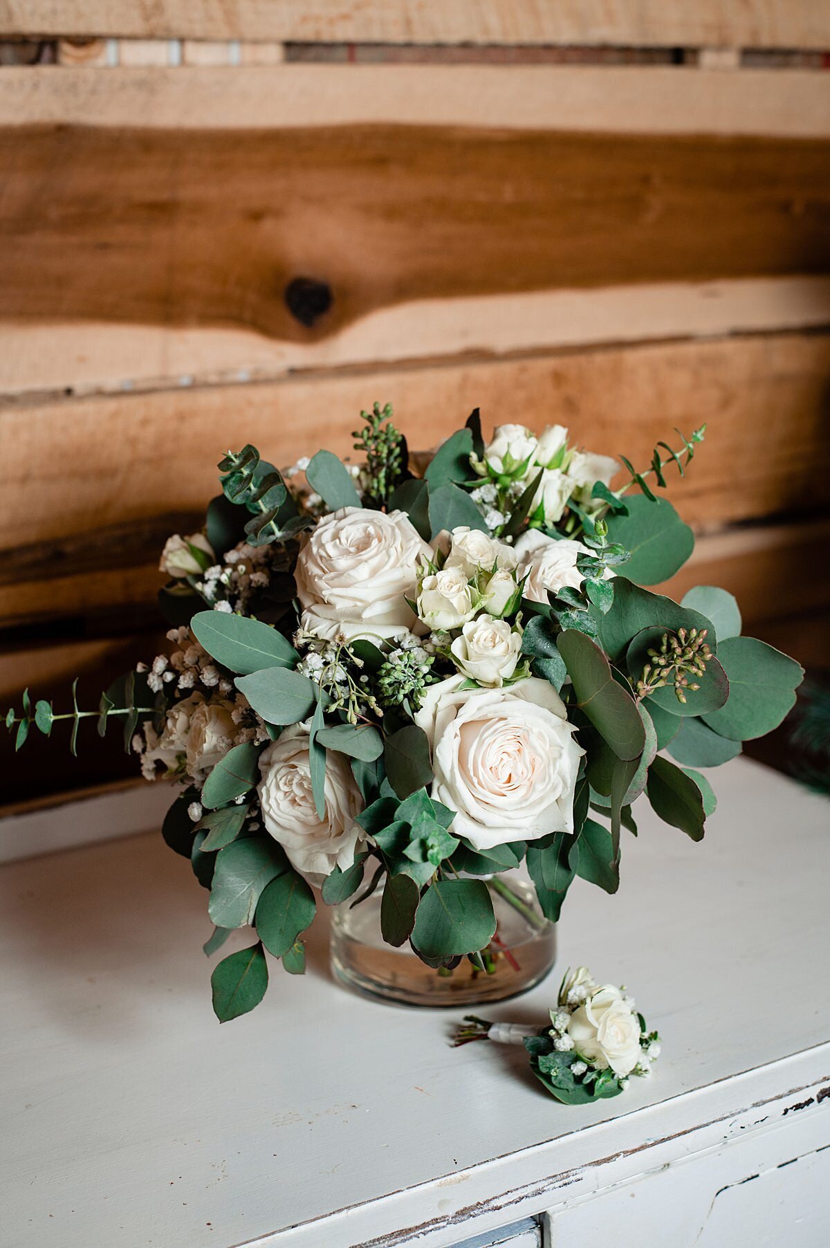 An elegant white bouquet of white roses, seeded eucalyptus, blue eucalyptus and spray roses in a glass vase sitting on a white farm table against a brown barn wall with a matching boutonniere.
