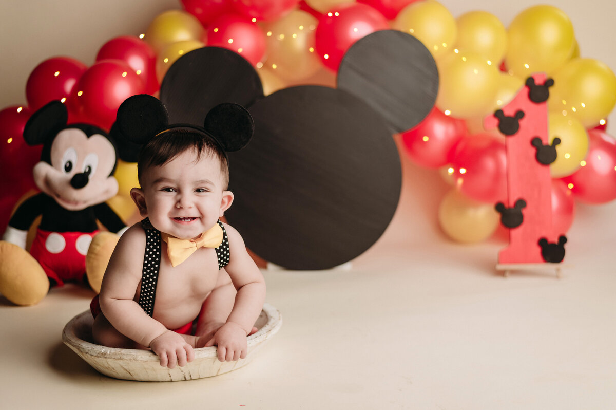 smiling baby boy wearing a mickey outfit  sitting on white wooden bowl in front of  large black mickey ears and white  and yellow balloons backdrop