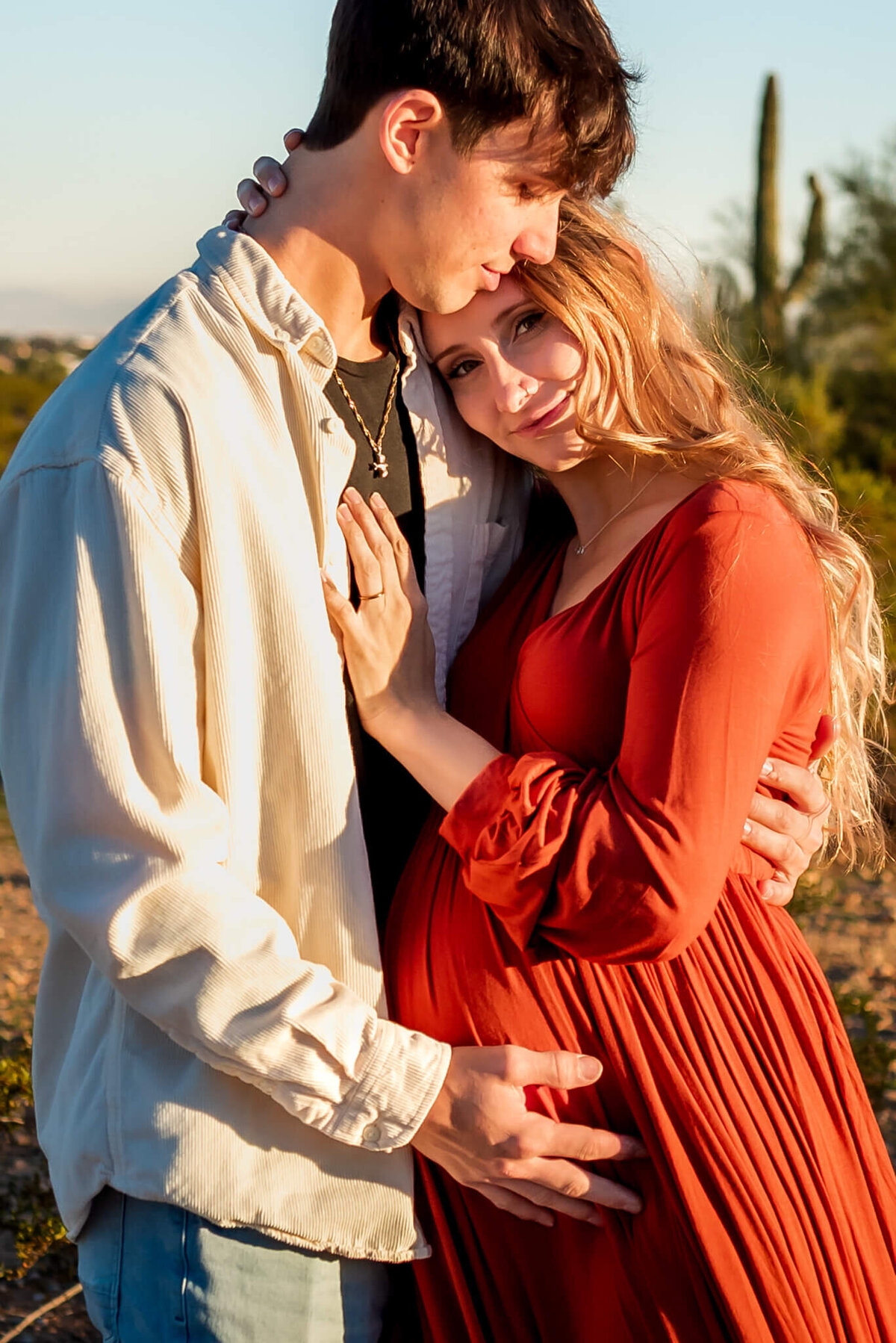 mom-to-be held in husband's embrace while he touches her baby bump. Photographed by Cactus & Pine Photography at late sunset in Arizona