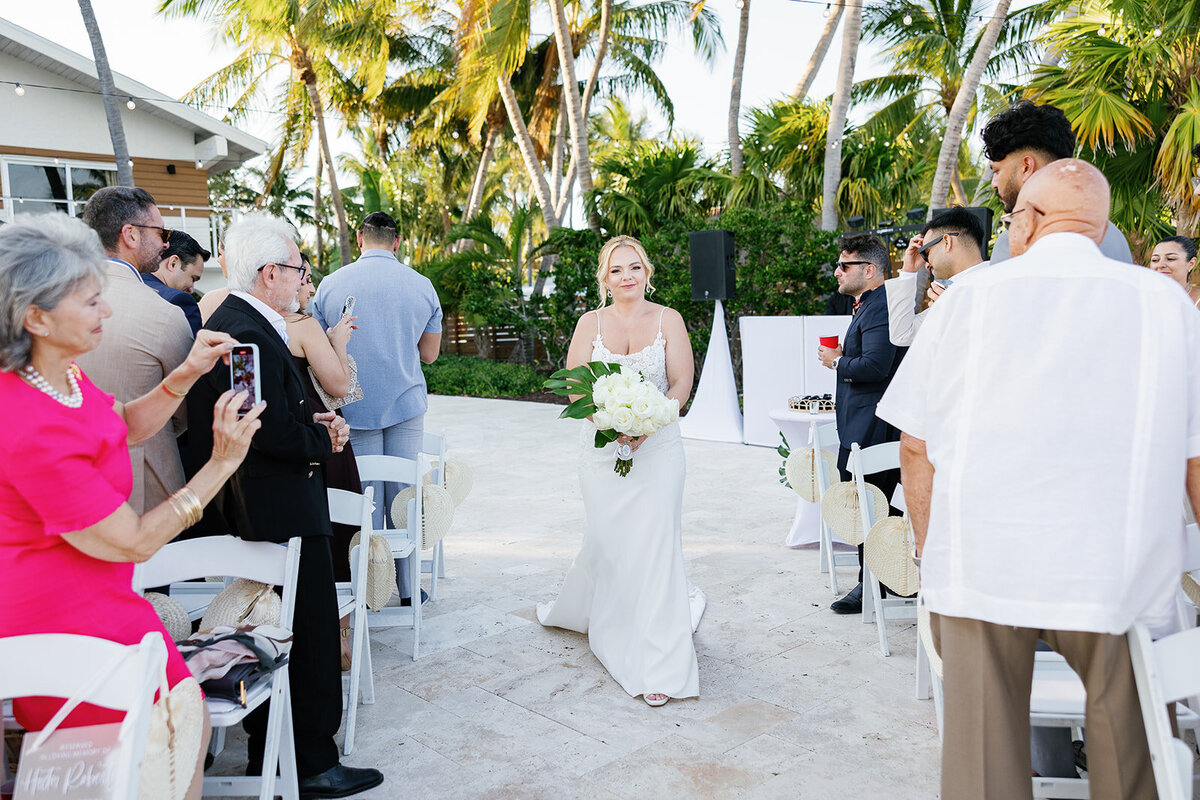 Kelly makes her way down the aisle holding a white bouquet, with guests capturing the moment at her Islamorada wedding.