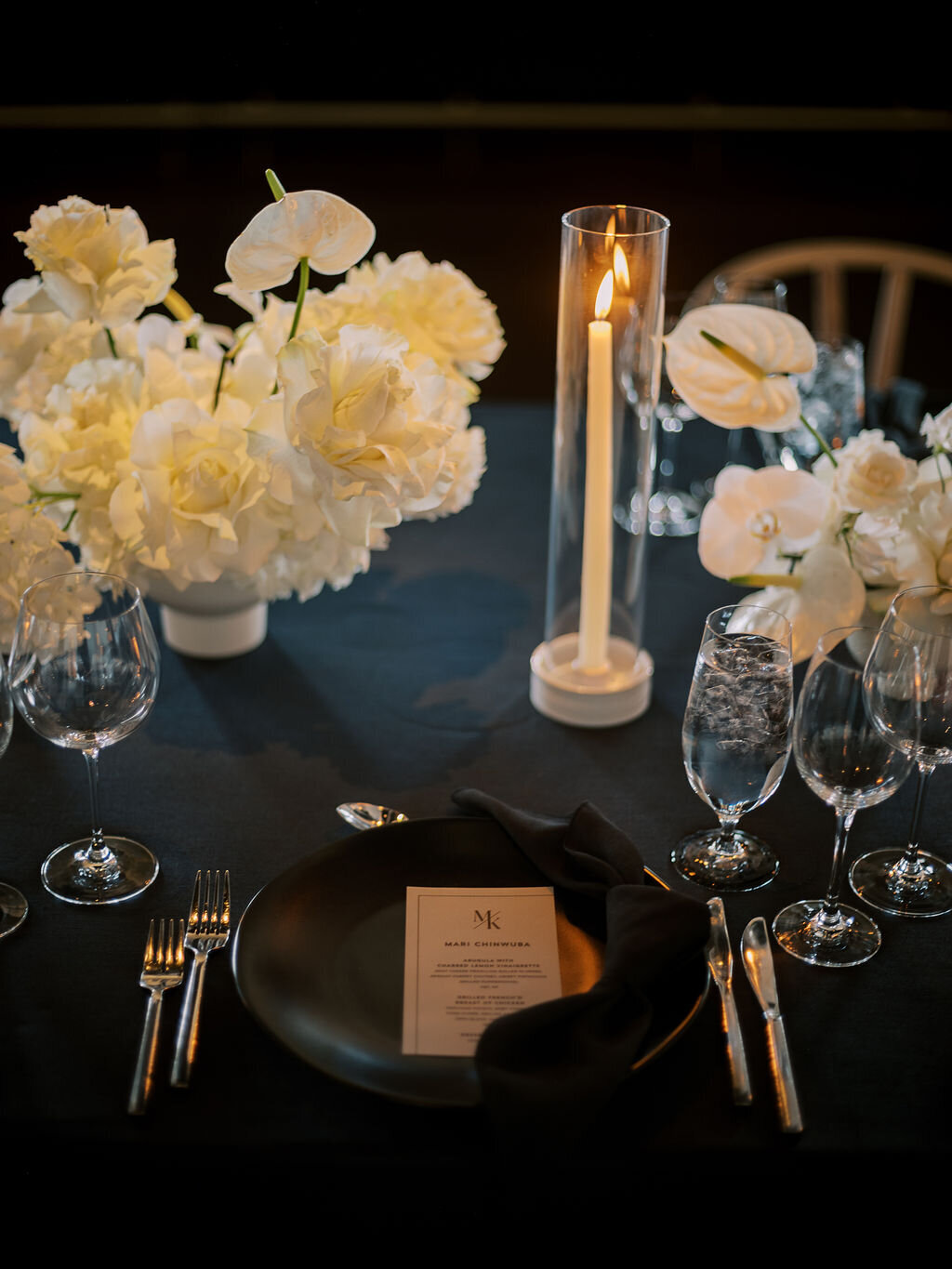 A dramatic reception table set with black tablecloths, white floral centerpieces, glowing candles, and fine glassware.