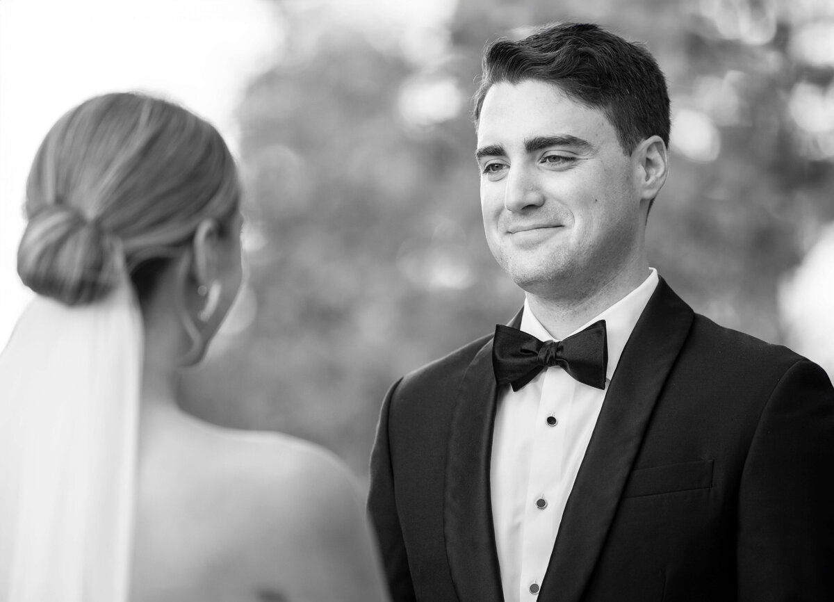 A groom in a tuxedo smiles at the bride, who is seen from behind. The background is softly blurred, highlighting the moments intimacy.