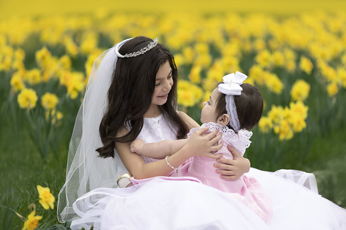 A young girl in a white dress sits in a field of daffodils playing with her toddler baby sister in a pink dress in her lap