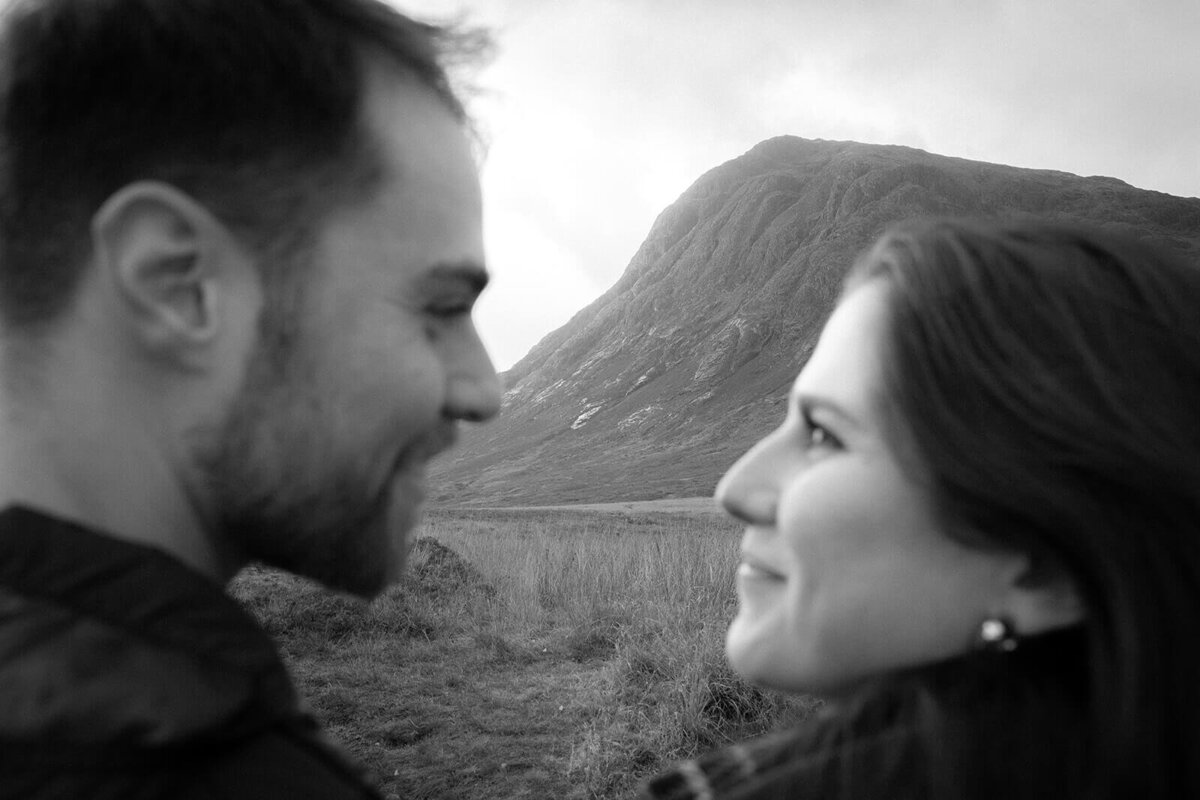The focus is on the mountain in the Scottish highlands as a couple in the foreground out of focus slightly stare lovingly at each other. 