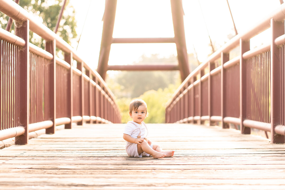 One-year-old boy wearing overalls sitting on a bridge looking at the camera