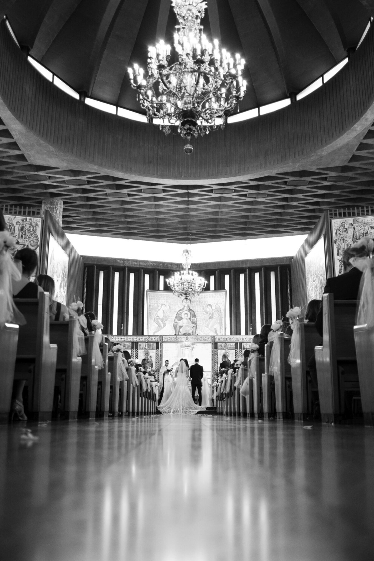 This black and white image captures the church aisle with the couple standing at the end, framed by the architectural beauty of the space. The monochrome tone highlights the elegance and solemnity of the wedding ceremony, focusing on the couple as they reach the end of their aisle journey.