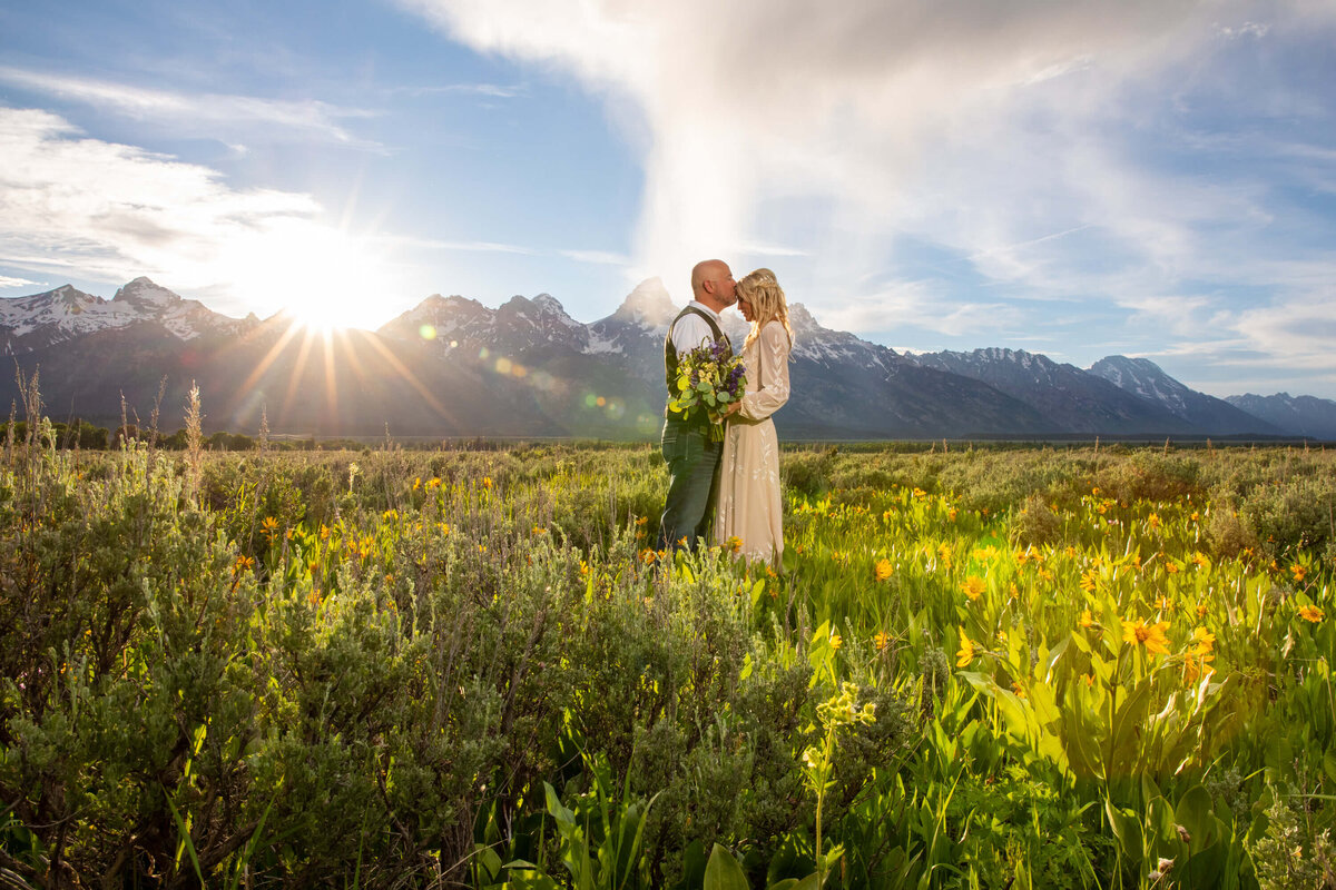 A bride and groom at sunset in a wildflower-filled meadow in Grand Teton National Park, with the sun casting a golden glow over the mountain peaks. The bride holds a bouquet of purple and yellow flowers.