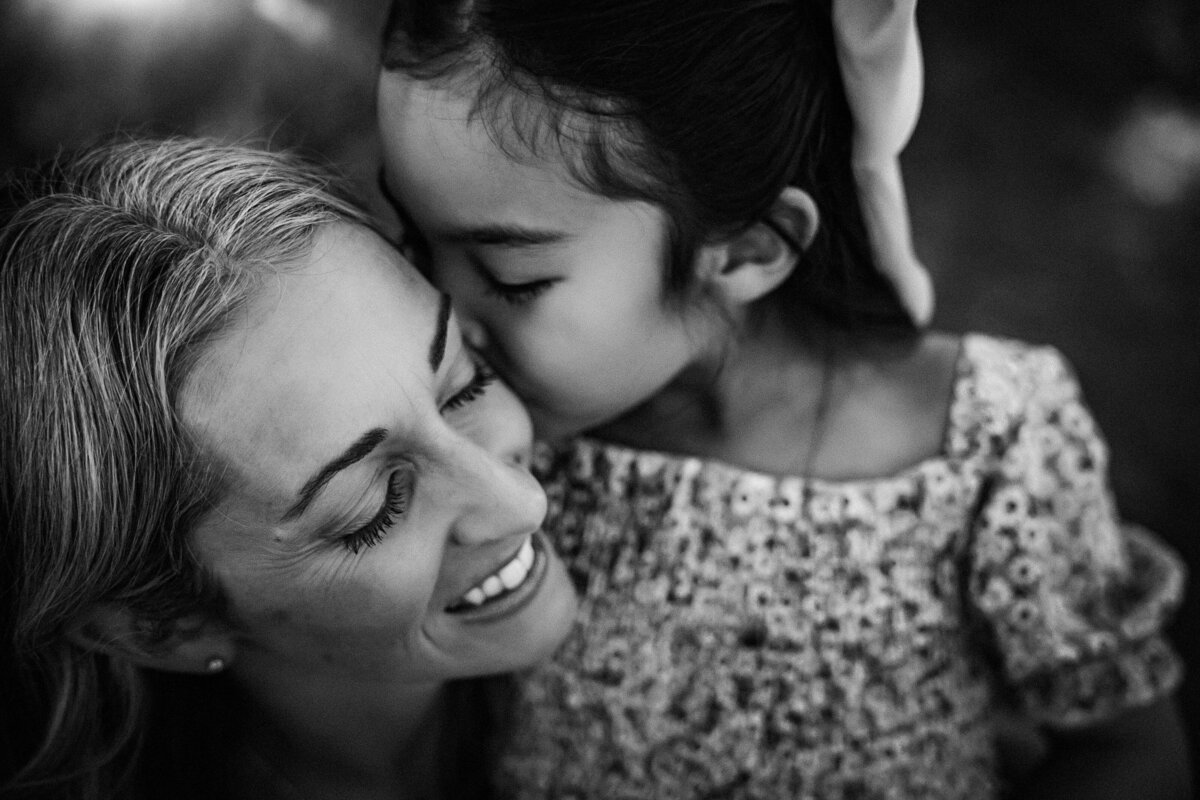 black and white image of girl kissing her mother's cheek. her mother is smiling and has her eyes closed.