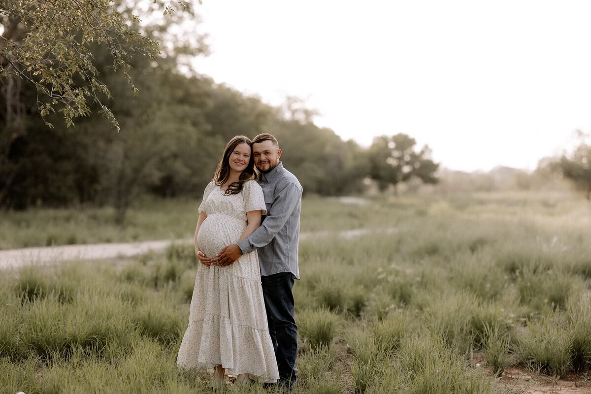 couple expecting their first baby, photo taken in front of a tree row with dads arms wrapped around mom