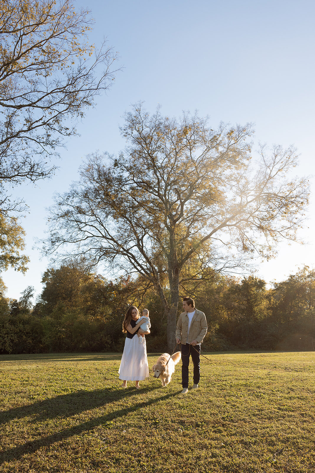 Family of three stands under large tree in the golden hour of sunset with their golden retriever by their side.