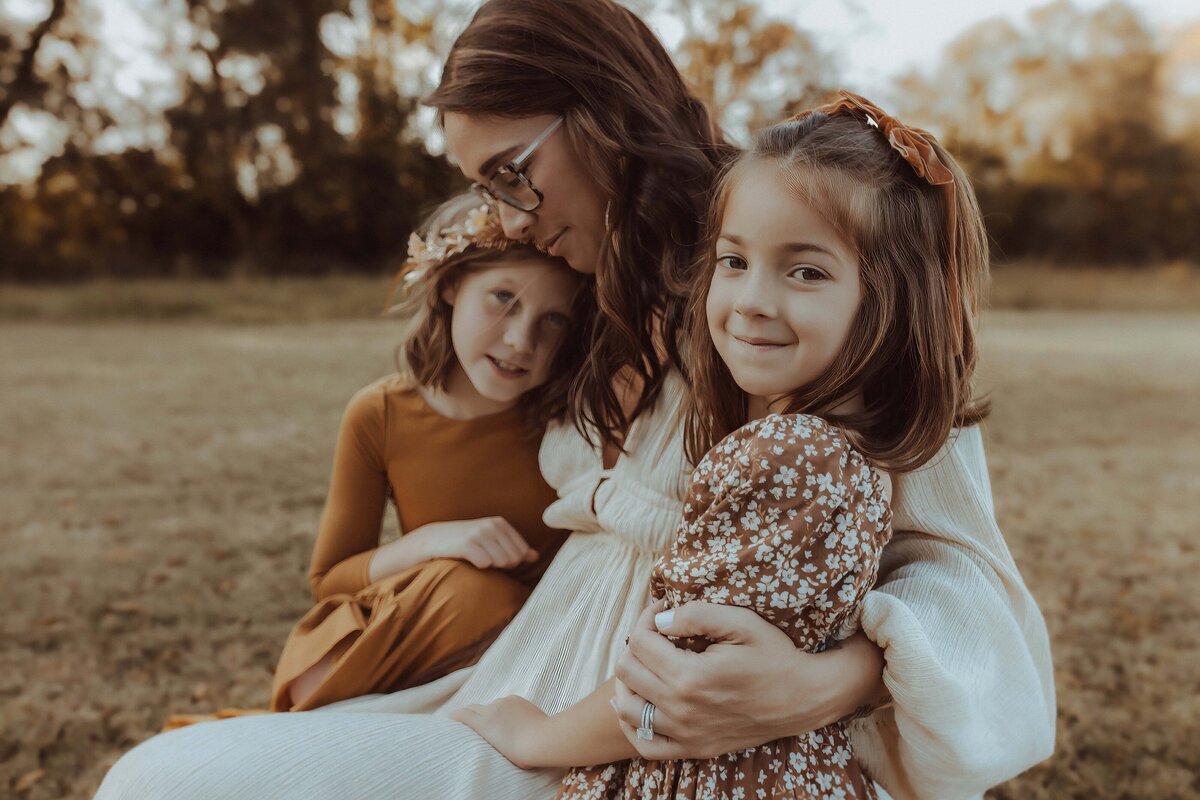 A woman in a white dress sits on grass with two young girls in floral dresses. One girl leans on her, and the other smiles at the camera. Trees are in the background.