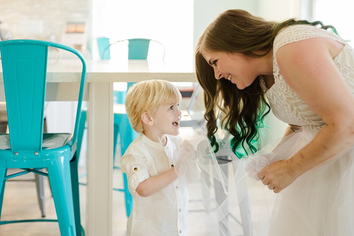 A bride leaning down to talk to her son before her wedding ceremony at Crystal Beach, Texas. The bride is wearing an intricate, white dress and is smiling down at her son. Her son is wearing all white and is happily holding her veil.