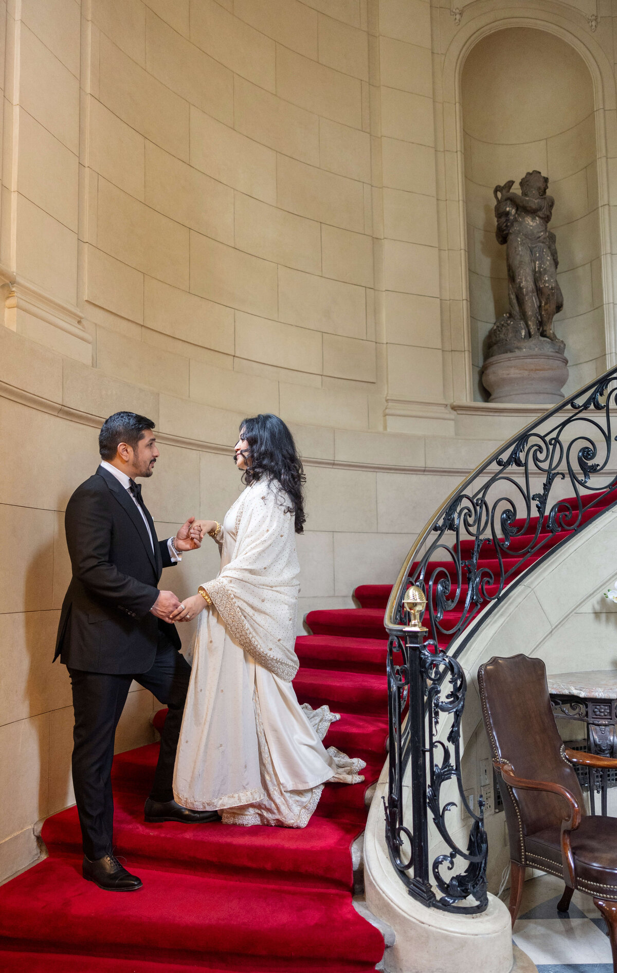 A couple in formal attire stands on a red-carpeted spiral staircase. The man wears a black suit, and the woman is in an elegant white gown. They hold hands, smiling, in a grand setting with a statue and ornate railing.