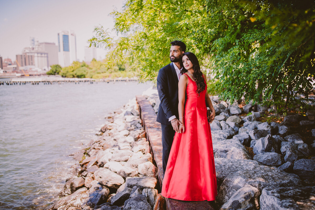 an engaged couple in red formal outfits standing on a jetty at the brooklyn bridge park in nyc