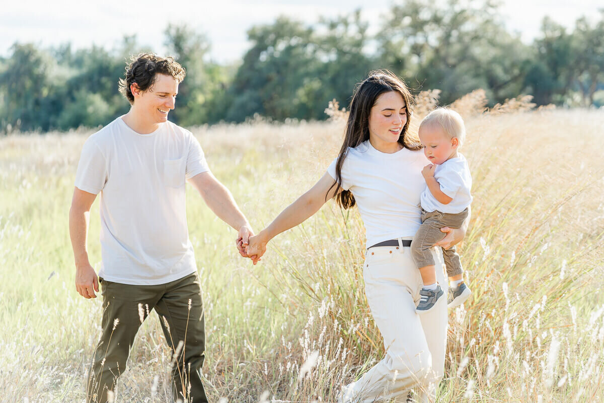 a mother holds her toddler son and pulls her husband's hand to walk through a field of tall grass during their fall family session in Austin, TX.