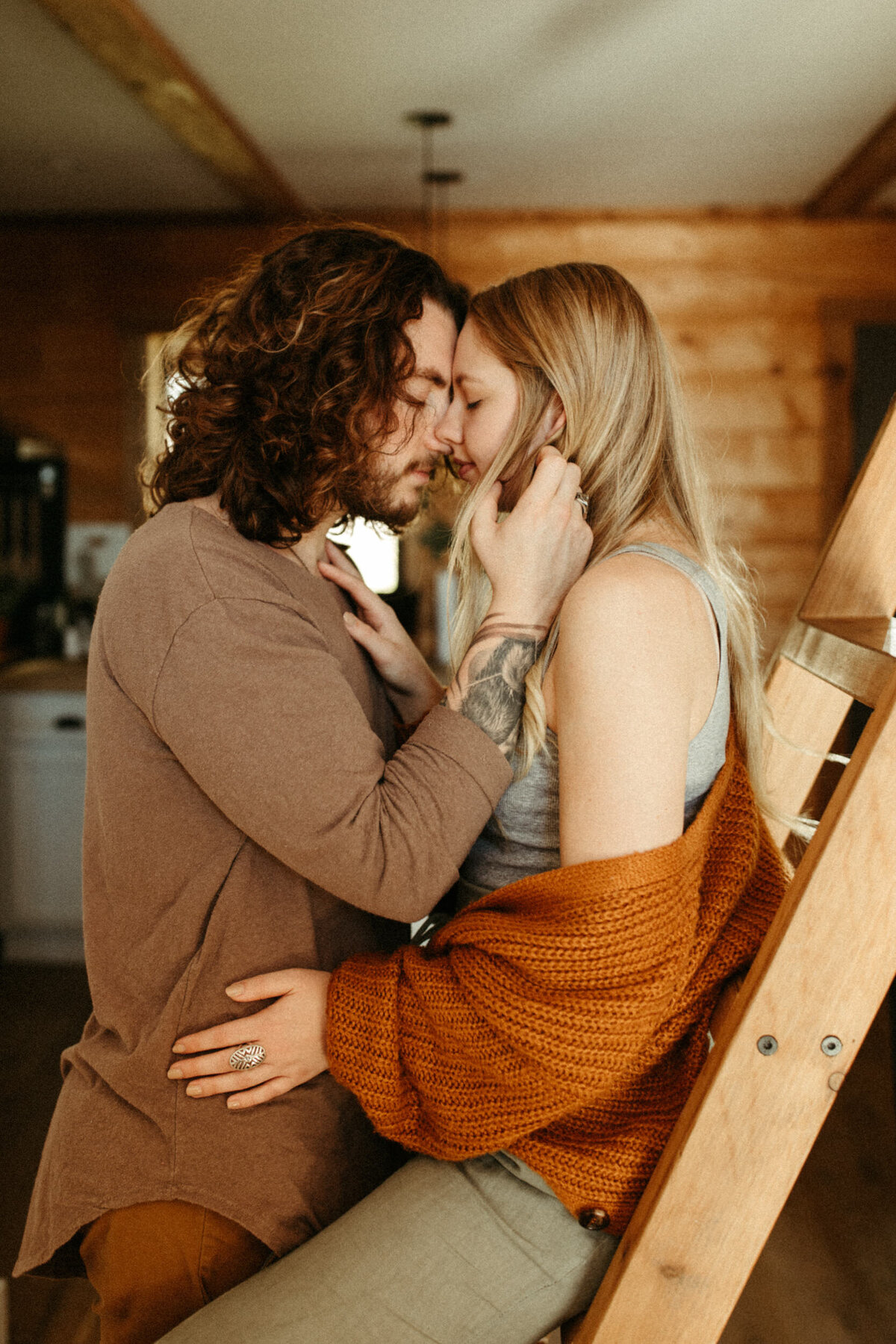 A girl is sitting on a wooden ladder inside a cabin while her fiancé is standing in front of her and pressing his forehead against hers.