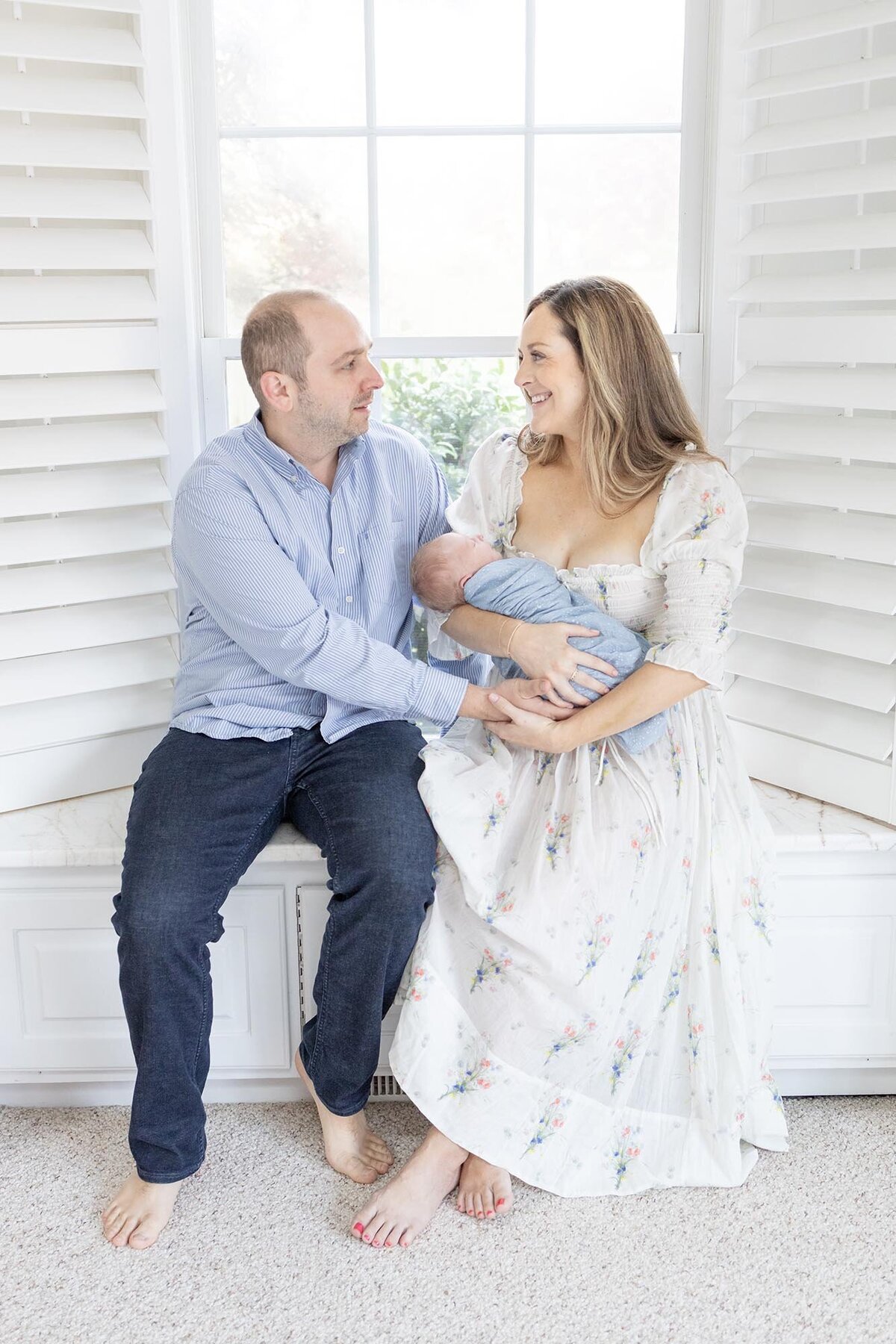 Mom and Dad sit on a window seat smiling at eachother snuggling their newborn baby by Virginia beach Newborn photographer Mary Eleanor