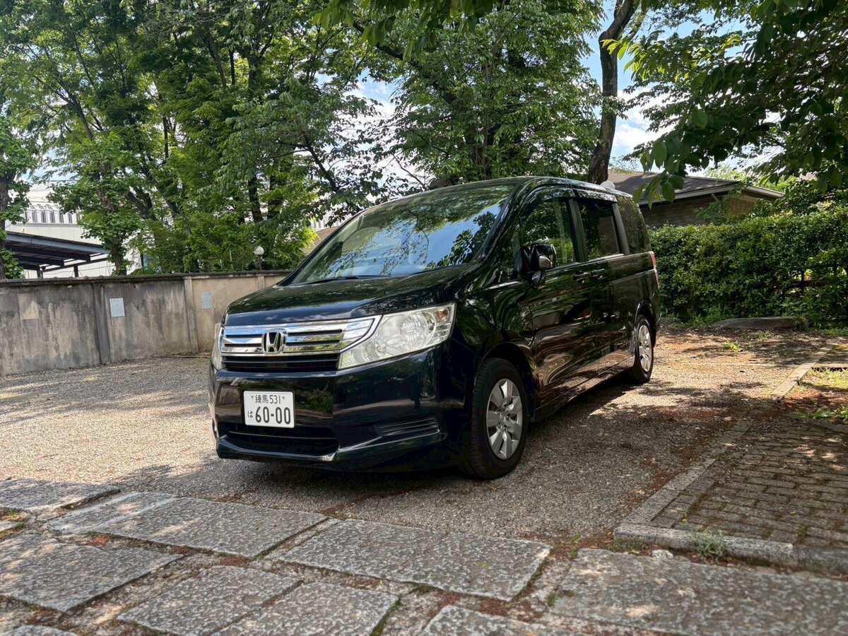A black Honda Stepwgn parked in a serene Japanese temple courtyard surrounded by lush greenery and traditional architecture under a sunny sky.