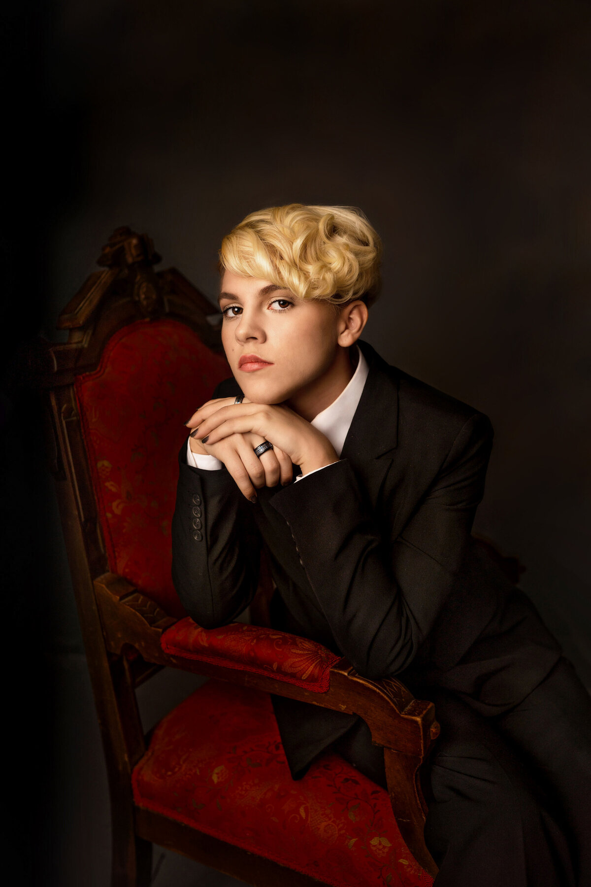 In studio image of female high school studio with pixie cut blonde hair.  She is wearing a black suit and sitting on a Victorian style wood chair with red upholstery.  Her hands are crossed  under her chin.  She has a serious expression.