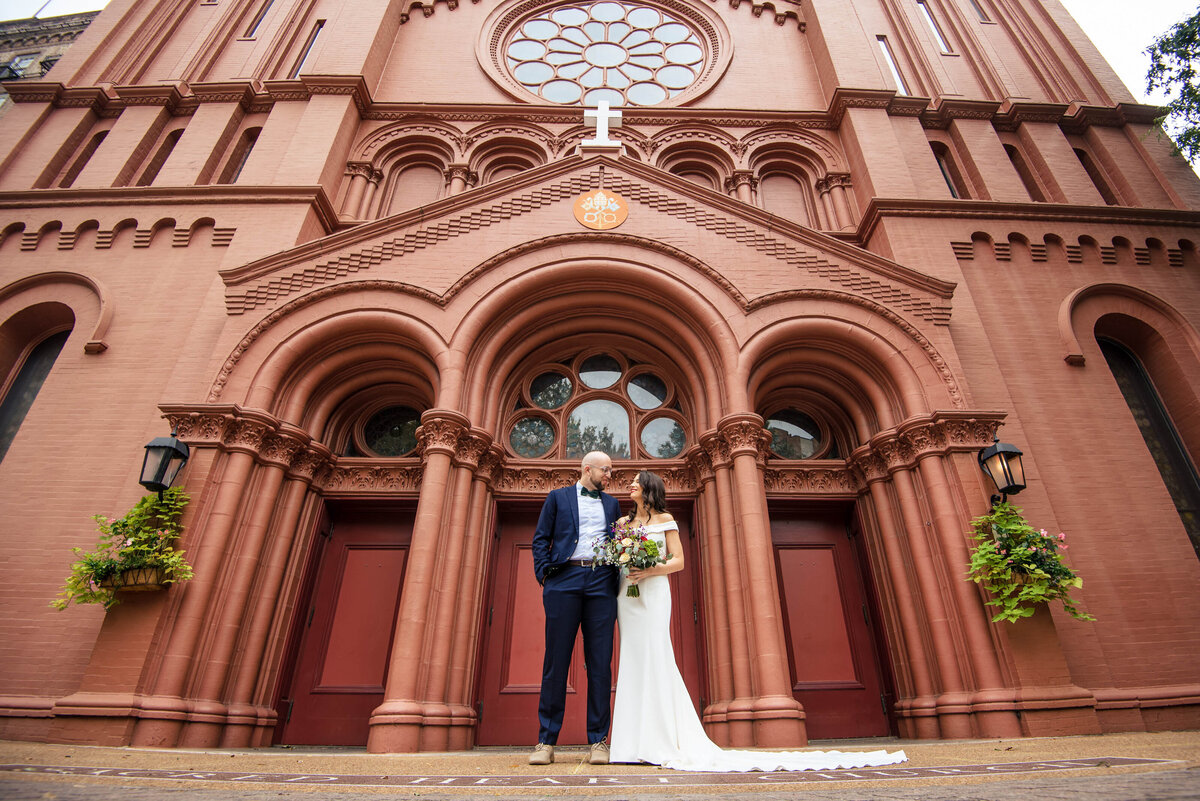 Low angle perspective photograph of bride and groom standing side-by-side smiling at one another outside Basilica of the Sacred Heart of Jesus church in Atlanta by Charlotte wedding photographers DeLong Photography