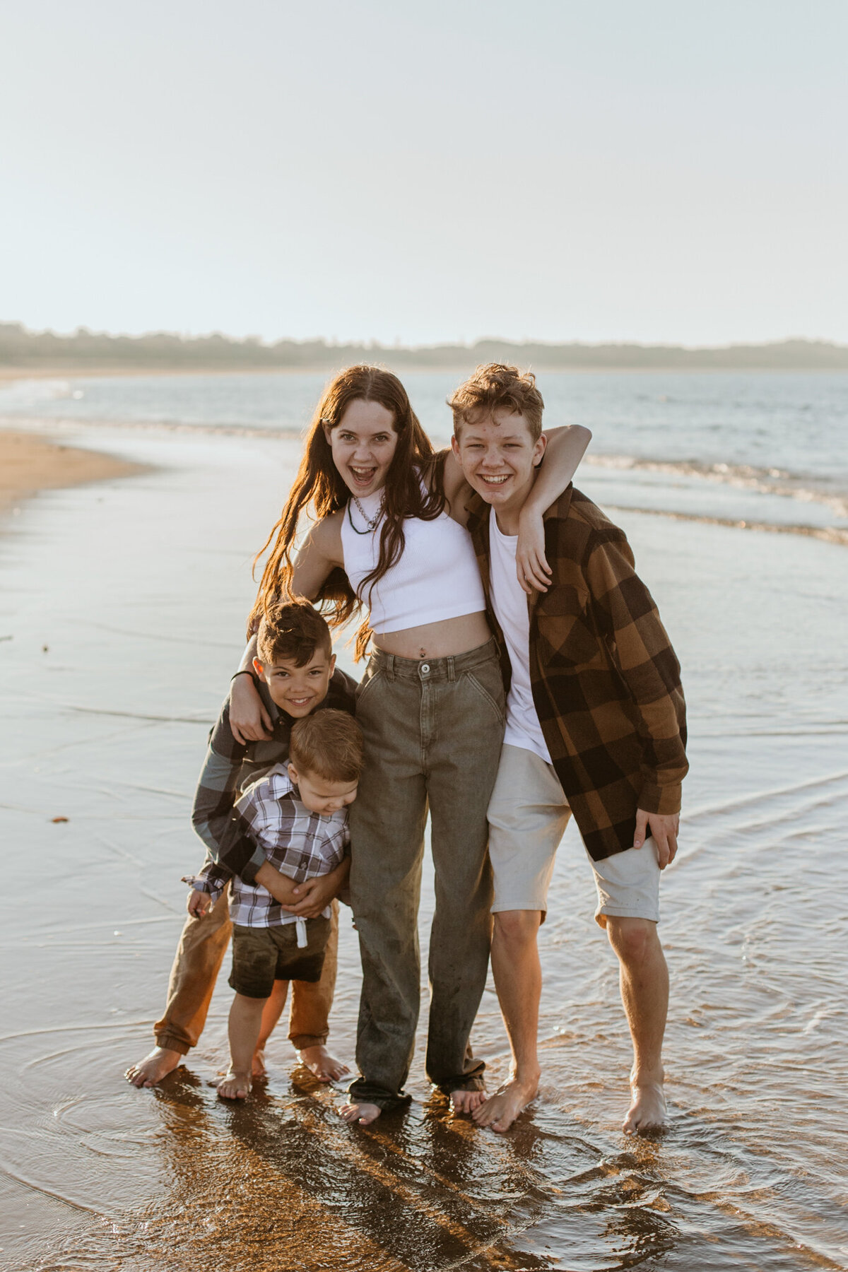 Four children share a joyful, spontaneous moment on the beach, captured in a documentary style, laughing and hugging in shallow water.