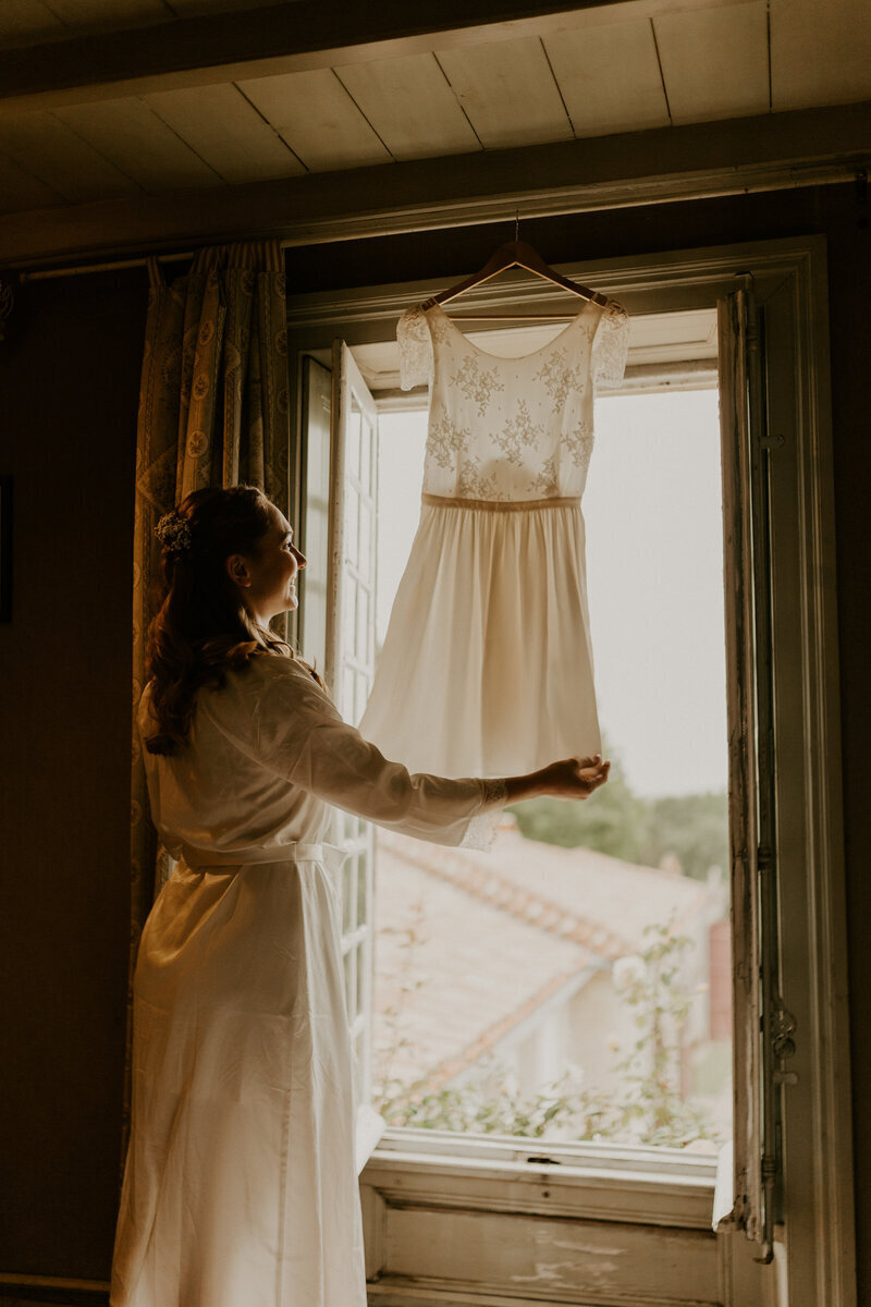 Mariée en peignoir tenant sa robe de mariée en dentelle blanche accrochée à un cintre à une fenêtre ouverte pour une séance photo mariage avec Laura, en vendée.